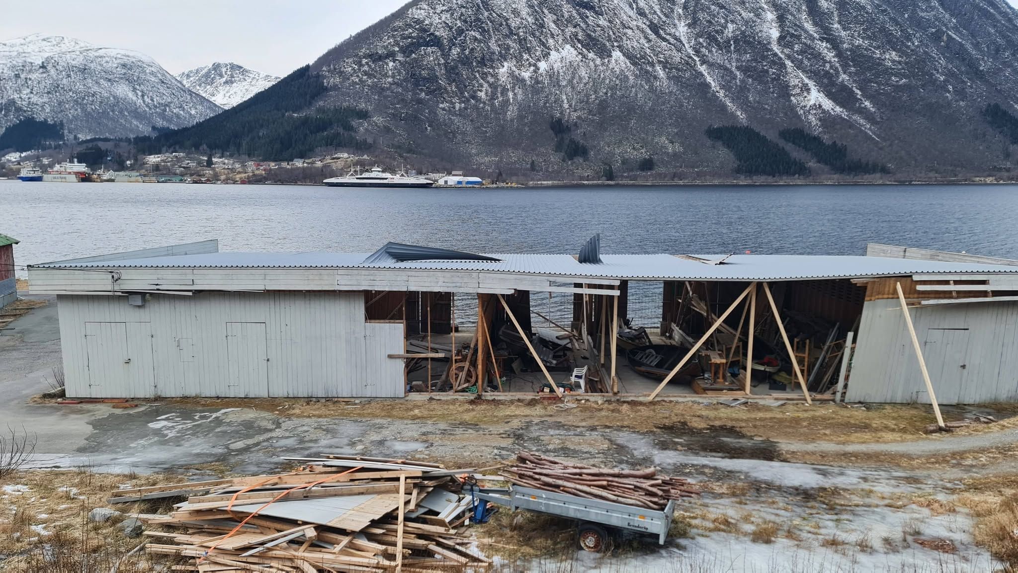 Eldre bygg ved sjøen der deler av veggkledningen er tatt ned og avstivet, med gamle plater og materialer sortert i en haug foran