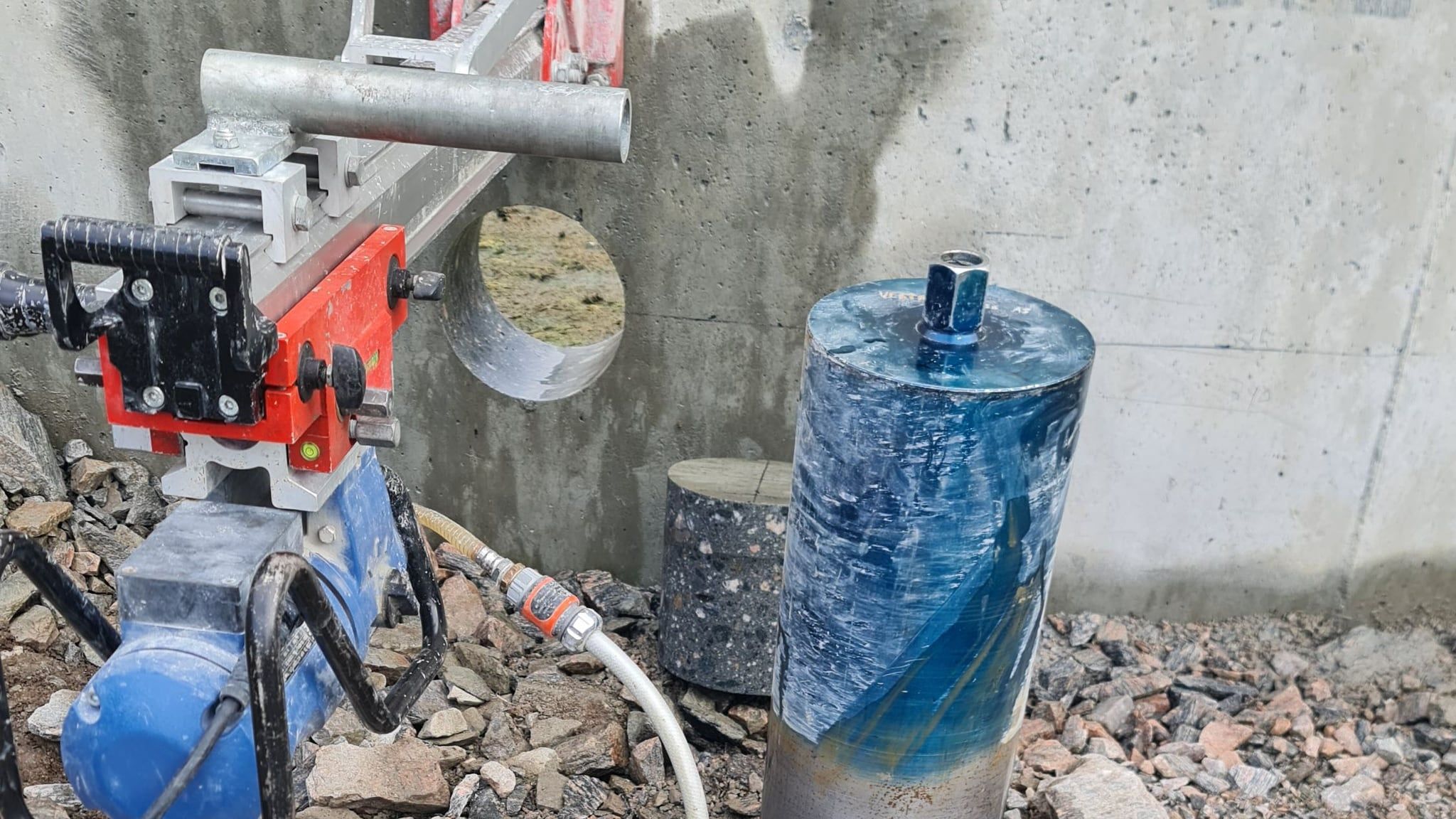 Borerigg montert mot en betongvegg med ferdig boret gjennomføring i veggen og den utborede betongkjernen stående på bakken ved siden av